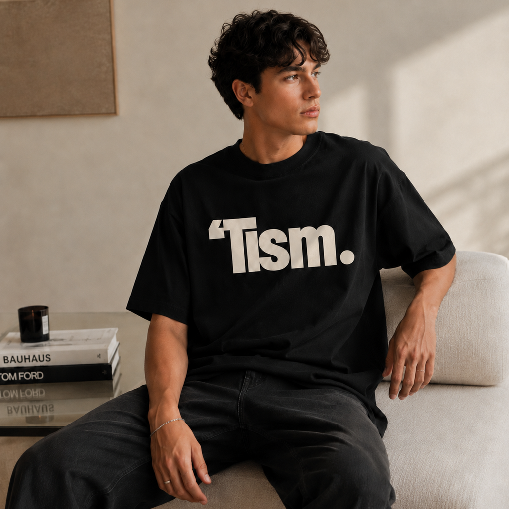 Person wearing a black t-shirt with 'Tism.' text, sitting on a couch in a minimalistic room.