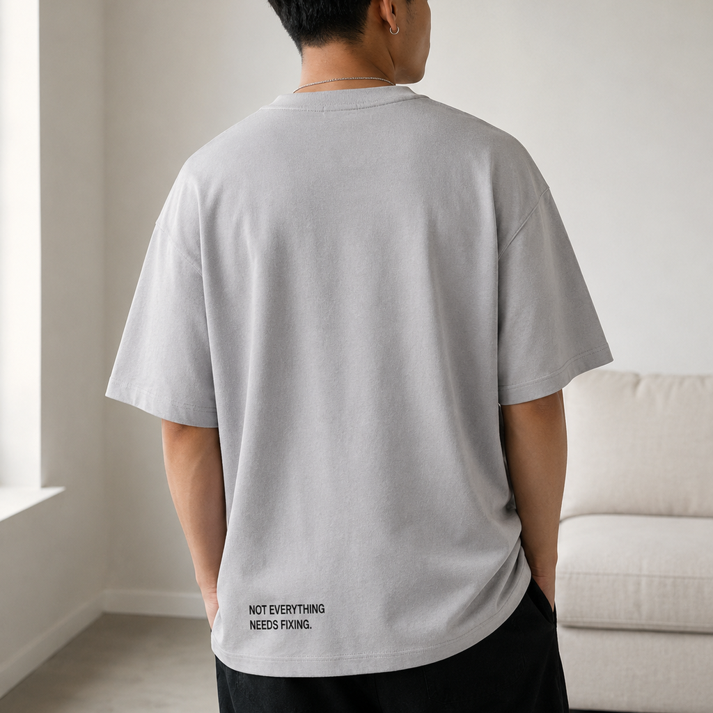 Person wearing a gray t-shirt with text on the back in a room with a white wall and beige sofa.