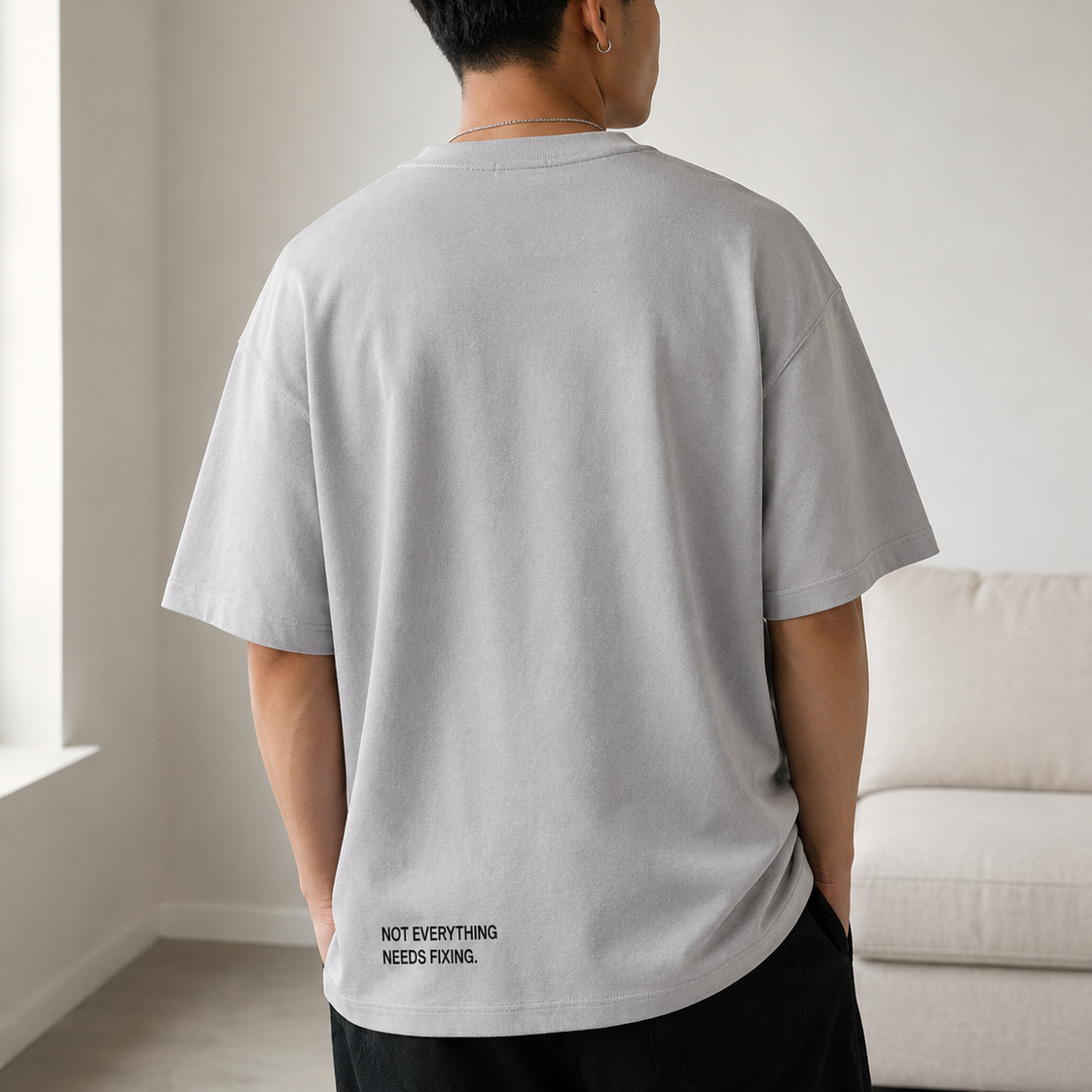 Person wearing a gray t-shirt with text on the back in a room with a white wall and beige sofa.