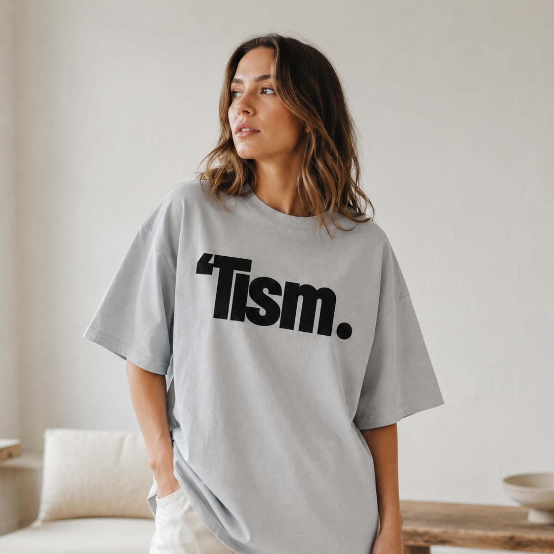 Woman wearing a gray t-shirt with 'Tism.' text in a casual indoor setting