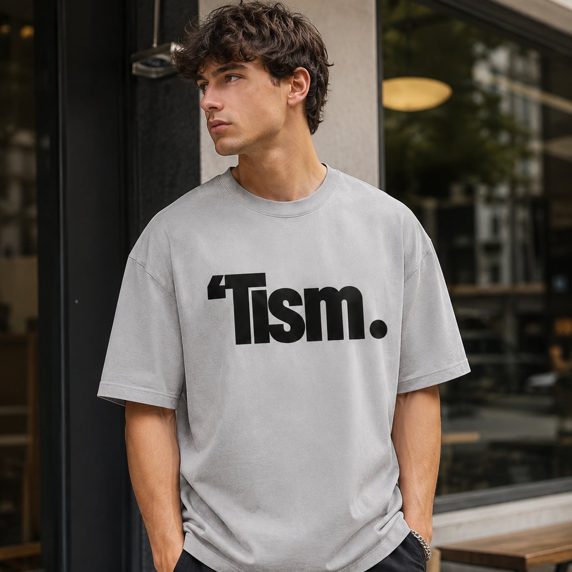 Person wearing a gray t-shirt with 'Tism' printed on it, standing outside a building.